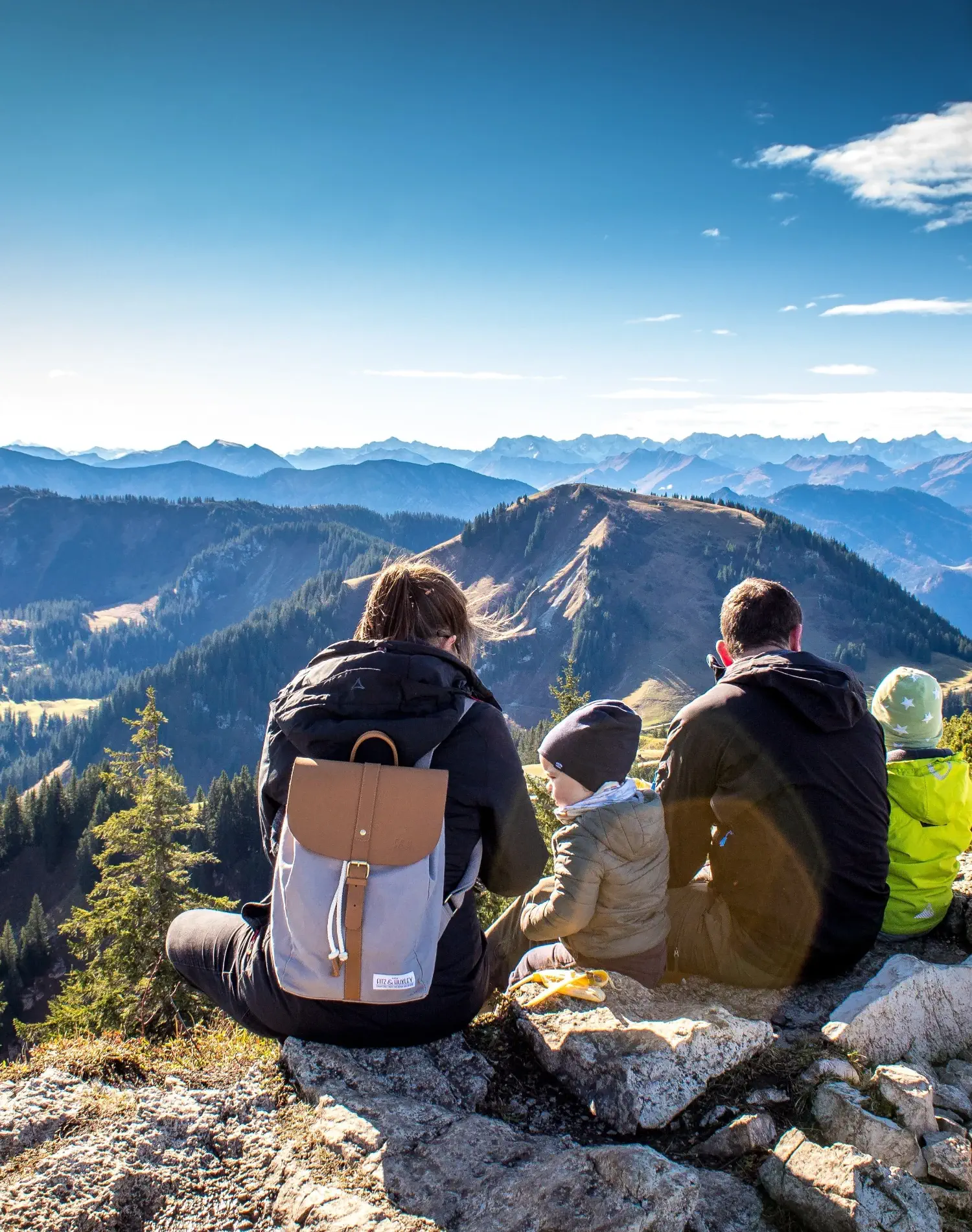 Eine vierköpfige Familie mit zwei kleinen Kindern, die auf einem Berggipfel sitzen. Von hinten fotografiert liegt vor ihnen ein tolles Bergpanorama bei strahlendem Wetter.